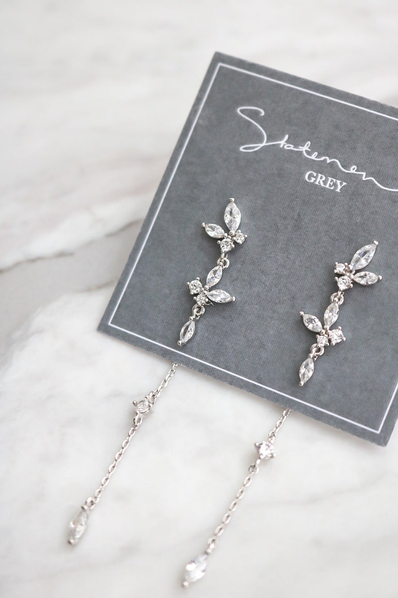 May include: A pair of silver dangle earrings with a delicate chain and a cluster of clear cubic zirconia stones in the shape of leaves. The earrings are displayed on a gray card with the text "Statement Grey".