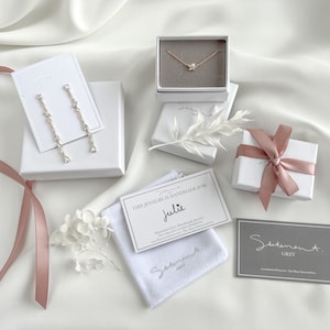 May include: A white gift box with a pink ribbon, a white jewelry box with a gold necklace, a white jewelry box with a pair of earrings, a white velvet pouch with a card that says "This jewelry is handmade for Julie", and a gray business card that says "Statement Grey".