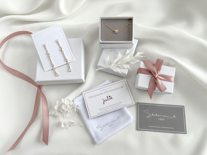 May include: A white gift box with a pink ribbon, a white jewelry box with a gold necklace, a white jewelry box with a pair of earrings, a white velvet pouch with a card that says "This jewelry is handmade for Julie", and a gray business card that says "Statement Grey".
