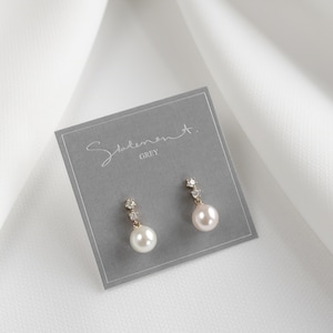 Puede incluir: Un par de pendientes de perlas con pequeños detalles de cristal en una tarjeta gris con el texto "Statement. GREY".
