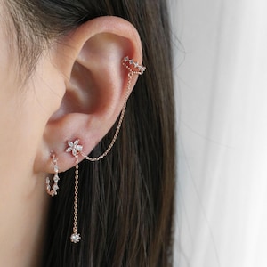 May include: Rose gold ear jewellery set. Includes a flower stud earring, a small hoop earring, and a chain that connects a flower stud to an ear cuff. The ear cuff and earrings are adorned with small, clear crystals.