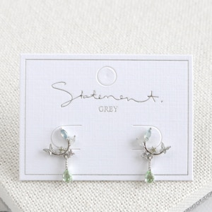 May include: Silver earrings with a delicate branch design, featuring light blue and green gemstones. The earrings are displayed on a white card with the text "Statement. GREY".