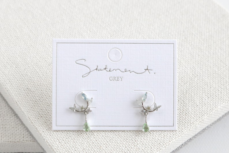 May include: Silver earrings with a delicate branch design, featuring light blue and green gemstones. The earrings are displayed on a white card with the text "Statement. GREY".