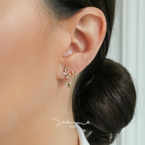 May include: Gold earrings with a leaf design and a dangling crystal charm. The earrings are worn in the left ear.