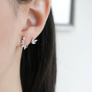 May include: A pair of gold earrings with a cluster of clear cubic zirconia stones in a leaf design. The earrings are shown on a model's ear.