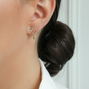 May include: A pair of gold earrings with a delicate leaf design and a dangling green gemstone. The earrings are worn in the left ear of a person with dark hair.