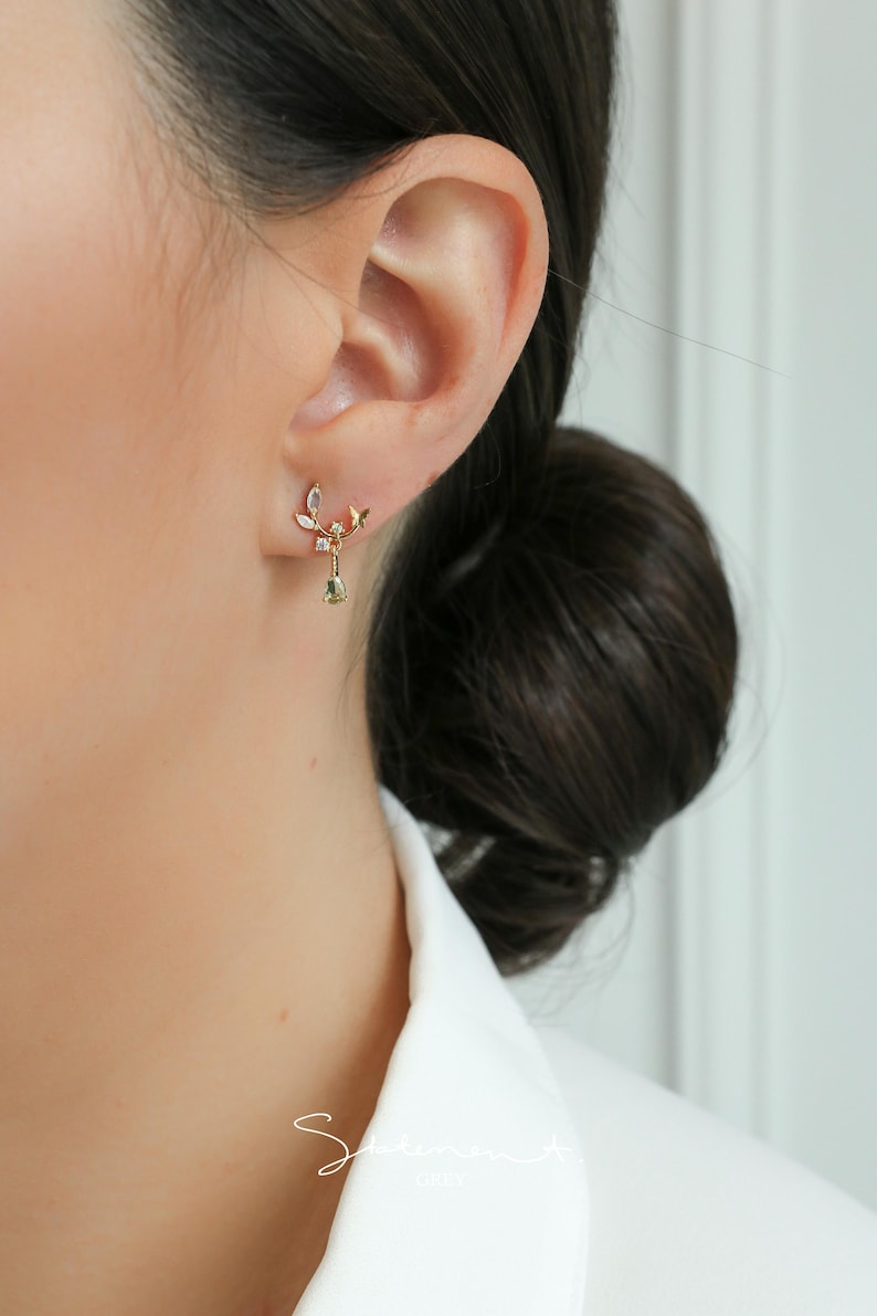 May include: A pair of gold earrings with a delicate leaf design and a dangling green gemstone. The earrings are worn in the left ear of a person with dark hair.