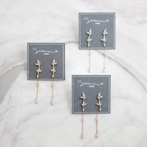 May include: Three pairs of dangle earrings with a floral design and a chain drop. The earrings are displayed on gray cards with the text "Sediment. Grey".