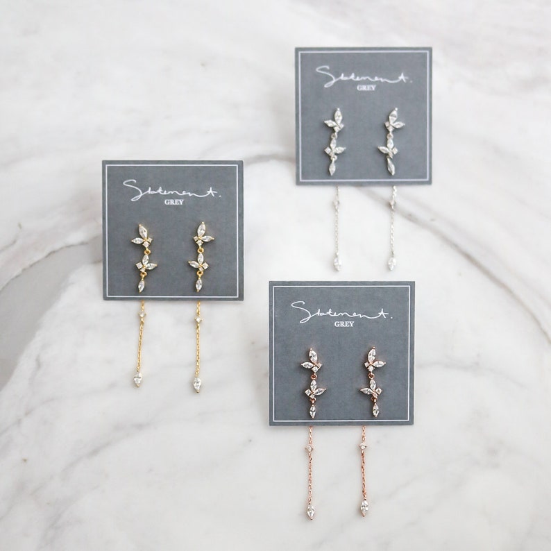 May include: Three pairs of dangle earrings with a floral design and a chain drop. The earrings are displayed on gray cards with the text "Sediment. Grey".