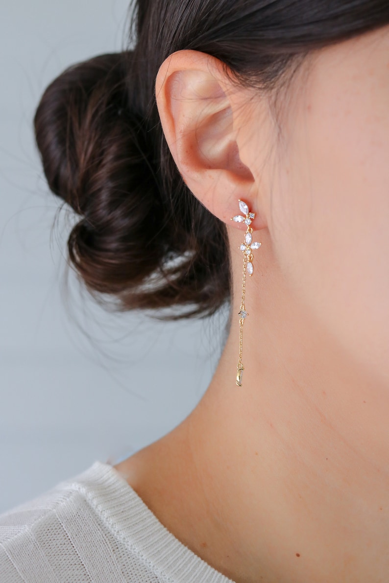 May include: Gold dangle earrings with a cluster of clear cubic zirconia stones at the top and a delicate chain extending down to a crescent moon charm.