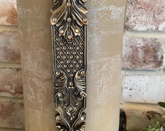 May include: A tall, cream-colored candle with a decorative gold metal handle. The handle is adorned with rhinestones and has a scroll-like design.
