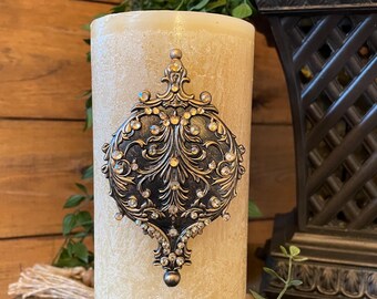 May include: A beige pillar candle with a decorative gold and crystal embellishment in the shape of a teardrop.