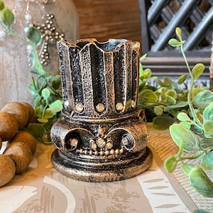May include: A decorative, antique-style candle holder. The holder is black and gold with a fluted design and embellished with rhinestones. The base has a crown design with more rhinestones. The candle holder is on a patterned surface.