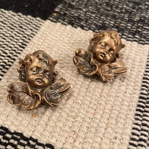 May include: Two gold cherub figurines with wings and a sleeping face. The cherubs are facing the same direction and are on a white and black checkered surface.