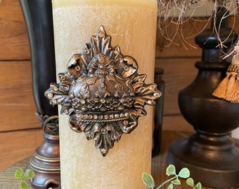May include: A tall, cream-colored candle with a textured surface. The candle is decorated with a large, ornate, silver-colored metal embellishment featuring a crown and intricate details. The candle is sitting on a black metal base with a decorative design.