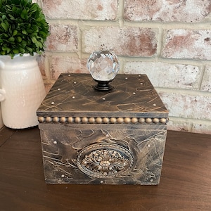 May include: A square wooden box with a distressed gold finish and a crystal knob. The box has a decorative metal design on the front and is adorned with small gold beads around the top edge.