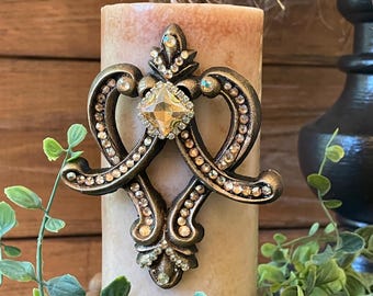May include: A decorative candle with a bronze-colored embellishment. The embellishment features a central, faceted, amber-colored gem surrounded by smaller, clear gems. The candle is beige with a mottled appearance.
