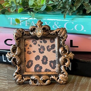 May include: A gold-toned ornate picture frame with a leopard print design inside. The frame has a decorative design with rhinestones on the corners.