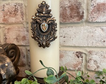 May include: A white pillar candle with a gold and crystal embellishment. The embellishment features a stylized design with a large crystal in the center.