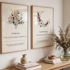 Puede incluir: Dos impresiones artísticas enmarcadas con diseños florales y de media luna. Las impresiones tienen texto en árabe e inglés. Un jarrón con flores secas y objetos decorativos se encuentra sobre una mesa de madera.