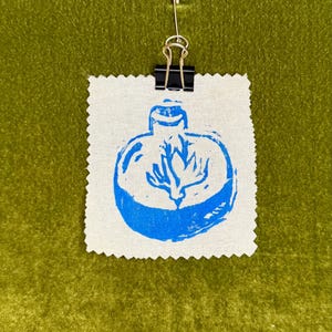 May include: A small, square fabric swatch with a blue print of a stylized ornament design. The fabric is held in place by a black binder clip and a silver safety pin. The background is a textured, olive green surface.