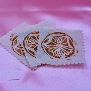 May include: Three small, off-white fabric squares with a brown, circular floral design. The edges of the squares are scalloped. The background is a solid, light pink fabric.
