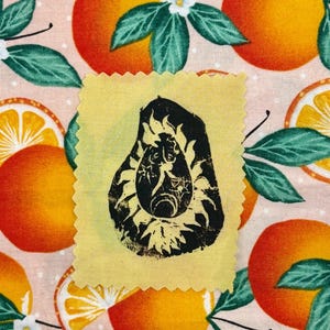 May include: A yellow fabric swatch with a black avocado-shaped design featuring a sun and moon motif. The swatch is set against a background of oranges, orange slices, green leaves, and white flowers on a pink dotted fabric.