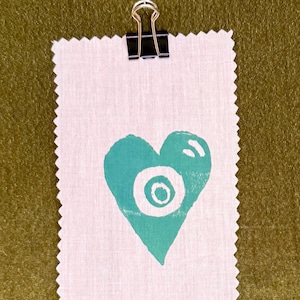 May include: A white fabric swatch with a teal heart design, held by a gold and black clip. The heart features a central circle and a surrounding ring. The fabric has a textured, raw edge and is set against a green background.