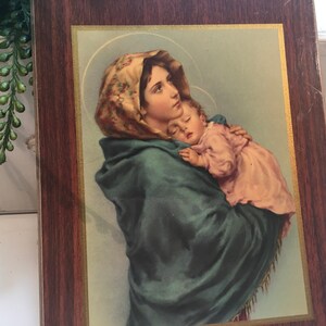 以下が含まれることがあります： 青いローブを着た女性が眠っている子供を抱いている宗教画の額縁。女性は花柄のヘッドカバーをしています。絵は薄い青色の背景です。