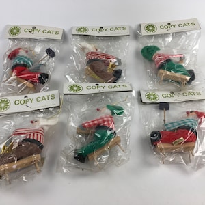 May include: Six vintage Christmas ornaments featuring Santa Claus figures in various poses, each wearing a red and white plaid shirt and a green hat. The ornaments are packaged in clear plastic bags with a white label that reads "COPY CATS".