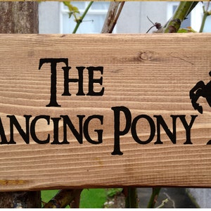 May include: A wooden sign with the text "The Prancing Pony" in black lettering. A black silhouette of a horse is on the right side of the sign.