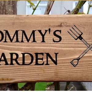 May include: A wooden sign with the text "TOMMY'S GARDEN" in black lettering. The sign features a crossed shovel and pitchfork in black.