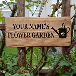 May include: A rustic wooden sign, reading "YOUR NAME'S FLOWER GARDEN" in black, with a black watering can illustration. The sign is mounted on a wooden fence, surrounded by lush green foliage and a glimpse of a house in the background.