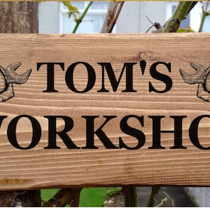 May include: Wooden sign with the text "TOM'S WORKSHOP" in black lettering. The sign is decorated with two stylised images of hands holding spanners.