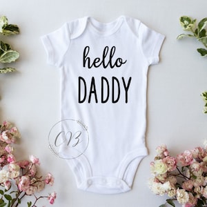 May include: White baby onesie with black text that reads "hello daddy".