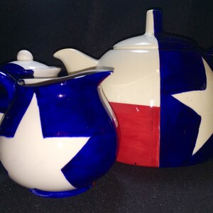 May include: A white teapot, creamer, and sugar bowl with blue and red accents. The teapot and creamer feature a white star on a blue background, while the sugar bowl has a blue star on a white background. The teapot and creamer are decorated with a red, white, and blue design reminiscent of the Texas state flag.