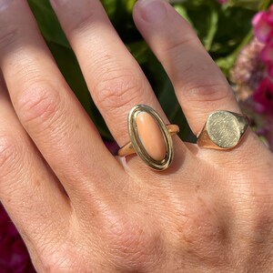 May include: A gold ring with a large oval-shaped pink coral stone set in a gold bezel. The ring is on a person's finger.