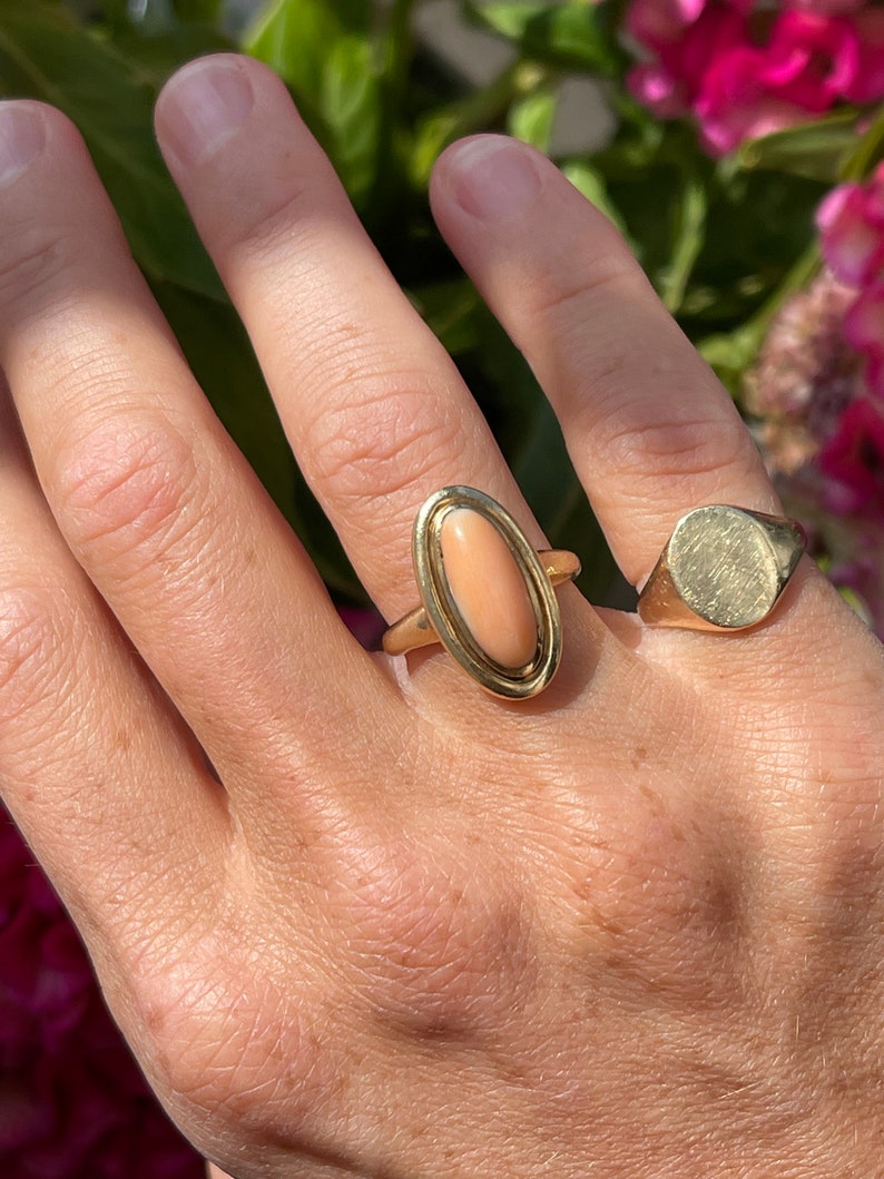 May include: A gold ring with a large oval-shaped pink coral stone set in a gold bezel. The ring is on a person's finger.