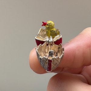 May include: A miniature Easter egg pendant featuring a yellow chick emerging from a red and silver egg. The chick has a red beak, and the egg's shell is silver. The egg is hinged, likely made of metal, and is a detailed piece of jewellery.