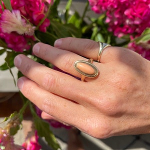 May include: A gold ring with an oval-shaped pink coral stone set in a gold bezel. The ring is on a person's finger.