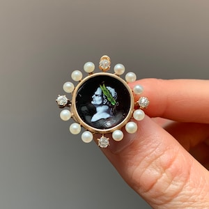 May include: A gold-toned brooch featuring a central black cameo portrait of a person with a laurel wreath. The cameo is surrounded by a ring of small pearls and accented with three sparkling diamonds. The brooch has a classic, elegant design.