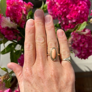 May include: A gold ring with a light pink oval gemstone. The ring is on a person's finger.