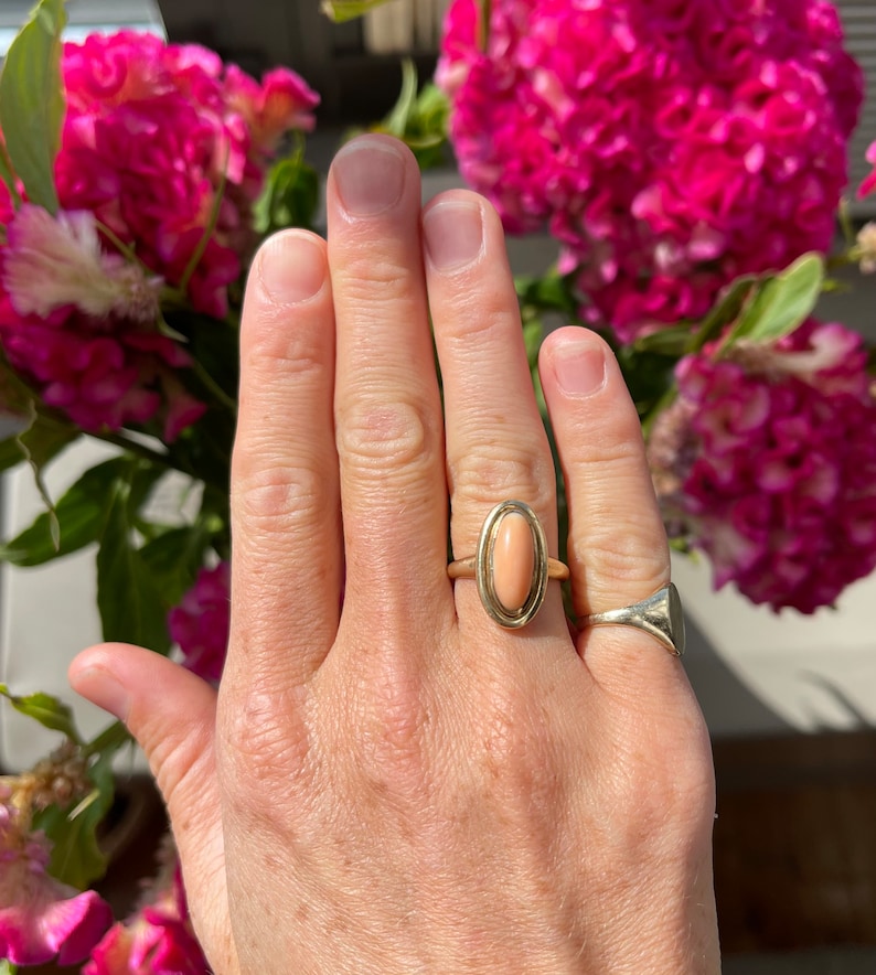 May include: A gold ring with a light pink oval gemstone. The ring is on a person's finger.