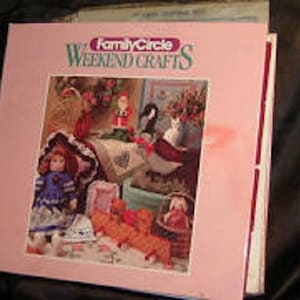 Pode incluir: Um livro rosa com o título "Family Circle Weekend Crafts" na capa. A capa apresenta uma boneca, uma cesta e uma variedade de outros materiais de artesanato.