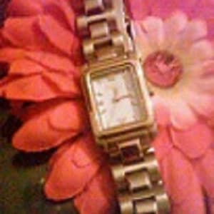 May include: A gold-toned wristwatch with a rectangular face and a linked band. The watch has a white face with gold hands and hour markers. The watch is resting on a background of pink and orange flower petals.