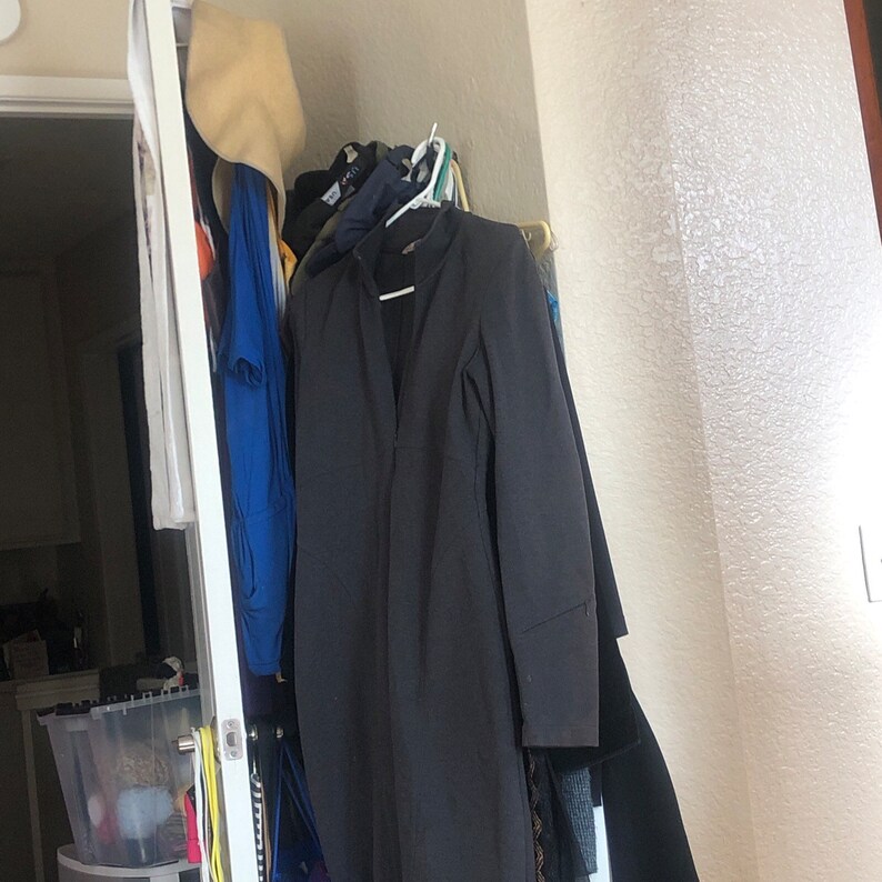 May include: A dark gray long-sleeved jacket hanging on a rack with other clothing, including a blue shirt and a straw hat.