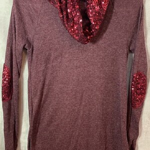 May include: A long-sleeved burgundy sweater with a cowl neck made of red sequins. The sweater has red sequin patches on the elbows.