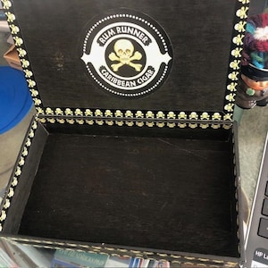 May include: A wooden box with a black background and gold trim. The lid has a skull and crossbones logo with the text "Rum Runner Caribbean Cigar".