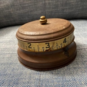 May include: A vintage, rusty, metal tape measure with a brass knob on top. The tape measure is marked with numbers 2, 3, and 4, and has markings for inches and centimeters.