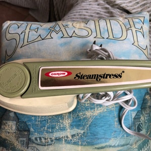 May include: A green and white handheld steamer with the brand name "Osrow" and the model name "Steamstress" printed on the front. The steamer has a white cord and a round, green button with the word "Caution" printed on it.